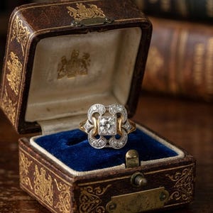 Pode incluir: Um anel de ouro com um diamante central de corte quadrado rodeado por uma moldura decorativa prateada com pequenos diamantes redondos. O anel é exibido dentro de uma caixa de joias de couro castanho com interior de veludo azul e detalhes dourados.