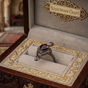 May include: A dark metal snake ring featuring a square-cut deep red gemstone set in gold. The snake body is textured with small dark stones and rests in a beige velvet-lined wooden box with gold embroidery and the text RAJASTHANI CRAFT.