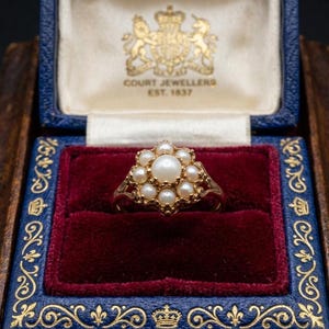 May include: A gold ring featuring a central pearl surrounded by a cluster of seven smaller pearls in a floral arrangement. The ring is presented inside a blue velvet-lined box with gold trim and the text COURT JEWELLERS EST. 1837 printed inside.