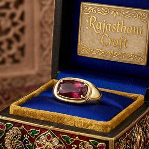 Peut inclure: Une bague en or avec une grande pierre précieuse rectangulaire rouge foncé montée dans un serti clos. La bague est présentée dans une boîte décorative avec une doublure en velours bleu et une plaque portant l'inscription Rajasthani Craft.