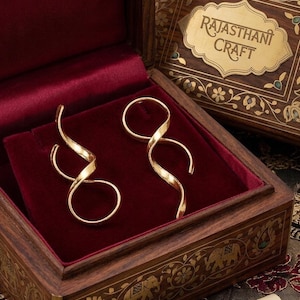 Puede incluir: Un par de pendientes en espiral de tono dorado con acabado pulido, presentados dentro de una caja de madera oscura forrada con terciopelo rojo intenso. La tapa tiene una incrustación ornamentada y una placa dorada con el texto RAJASTHANI CRAFT.