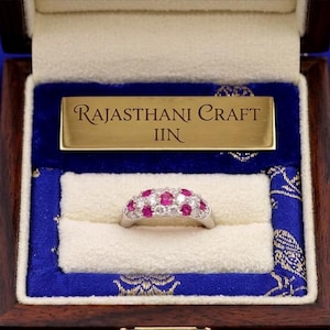 Può includere: Un anello color argento con un gruppo di pietre rotonde rosa e trasparenti, presentato in una scatola di legno scuro con una placca dorata incisa con il testo RAJASTHANI CRAFT IIN, su una fodera in tessuto blu con motivo a pavone.