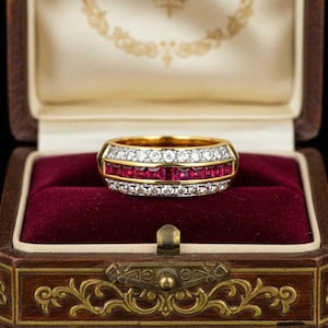 Könnte beinhalten: Ein Goldring mit einer mittleren Reihe aus quadratisch geschliffenen roten Rubinen, flankiert von zwei äußeren Reihen aus rund geschliffenen weißen Diamanten. Der Ring liegt in einer braunen Schmuckschatulle mit cremefarbenem Deckel und rotem Samt.