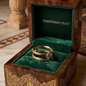 Può includere: Un anello d'oro a forma di serpente avvolto all'interno di una scatola di legno lucido con interno in velluto verde scuro. Il coperchio presenta la scritta dorata RAJASTHANI CRAFT e l'esterno è decorato con intricati motivi dorati.