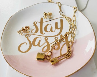 May include: Gold tone jewelry set with a chain necklace, a padlock charm, and a pair of star-shaped earrings. The jewelry is displayed on a white and pink ceramic dish with the words "stay sassy" written in gold.