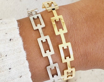 May include: Two gold and silver chain bracelets with a square link design. The bracelets are stacked on a person's wrist.