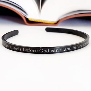 May include: A black metal bangle bracelet with the inscription "Only who kneels before God can stand before anyone".