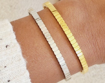 May include: Two minimalist bracelets, one gold and one silver, on a person's wrist. The bracelets are made of small, square-shaped links.