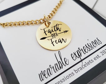 May include: Gold necklace with a round pendant that says "Faith over Fear" with an arrow pointing to the right. The necklace is in a box that says "wearable expressions" and "expressions bracelets est. 2017".