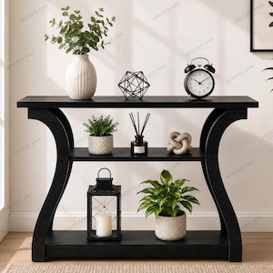 May include: A black wooden console table featuring a curved frame design with three horizontal shelves. The table is decorated with a white vase, a geometric wire sculpture, a black alarm clock, potted plants, a reed diffuser, and a lantern.
