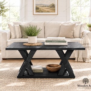 May include: A rectangular black wooden coffee table with an X-shaped base and a lower shelf. The tabletop holds a potted green plant on a woven mat and three stacked books titled The Kinfolk Home, Plenty, and Half Baked Harvest.