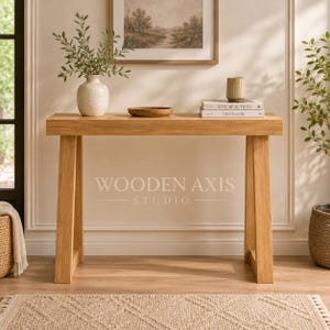 May include: A light brown wooden console table with A-frame legs. On the table sits a white textured vase with green foliage, a small wooden bowl, and two stacked books with the text "LIVE BEAUTIFUL" and "THIS IS HOME" printed on the covers.