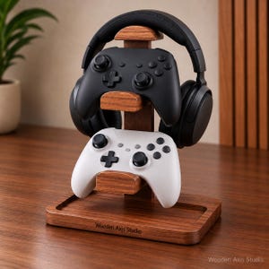 May include: A wooden stand featuring a top arch for headphones and two lower horizontal supports for game controllers. The base is rectangular with a recessed tray and the text Wooden Axis Studio printed on the front. A black controller and a white controller are resting on the supports.