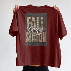 Könnte beinhalten: Ein bordeauxrotes Kurzarm-T-Shirt mit einer rechteckigen Grafik auf der Rückseite. Die Grafik zeigt den Text FALL SEASON in Blockbuchstaben mit verschiedenen Mustern wie Leopardenmuster, Karos und Punkten sowie das Wort cozy in Schreibschrift.