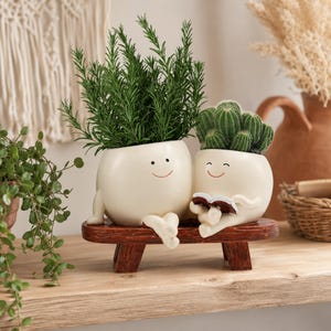 Peut inclure: Deux cache-pots en céramique crème, en forme de personnages souriants, assis sur un banc en bois marron. Le plus grand contient un romarin vert, tandis que le plus petit contient un cactus vert et tient un petit livre ouvert entre ses mains.