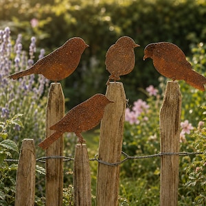 Könnte beinhalten: Vier rostfarbene Metallvogel-Silhouetten, die auf verwitterten Holzzaunpfosten sitzen. Jede Vogelfigur weist eine texturierte, oxidierte Oberfläche auf und ist sicher auf der Spitze eines einzelnen vertikalen Holzpfahls in einem Garten befestigt.