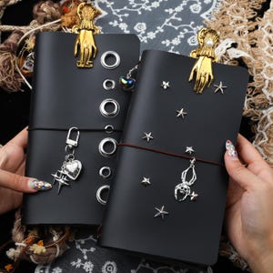 Könnte beinhalten: Zwei schwarze Notizbücher in Lederoptik mit goldenen Hand-Clips an der Oberseite. Das linke Notizbuch hat silberne Ösen und einen Herzanhänger. Das rechte ist mit silbernen Sternnieten und einem silbernen Spinnenanhänger mit Totenkopf verziert.