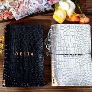 May include: Two rectangular journals with a textured crocodile skin pattern. The left journal is black with the name DELIA in gold letters. The right journal is silver with the name LANVJ in silver letters. Both feature elastic bands and charms.