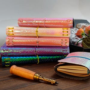 May include: A stack of five rectangular journals with iridescent, snakeskin-textured covers in pink, peach, coral, purple, and blue, each secured with coloured elastic cords and metal eyelets, alongside a wooden-handled tool and an open journal.