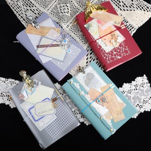 May include: Four rectangular journals in lavender, red, grey, and teal, each decorated with layered paper ephemera, lace, metal charms including hands and stars, and secured with coloured elastic bands. The covers feature various collage elements.