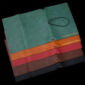 May include: A stack of five rectangular faux leather document folders in teal, tan, red, brown, and black. Each folder features a metal eyelet with a coloured elastic cord closure. The teal folder reads THE DISCIPLE FILES in gold text.