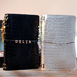 May include: Two rectangular journals with a glossy crocodile-embossed texture. The left journal is black with the name DELIA in silver letters. The right journal is silver with metallic letters. Both feature decorative charms and elastic bands.