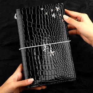 May include: A black journal with a glossy crocodile-embossed texture. The cover features silver star-shaped studs in the upper right corner. A white elastic band wraps around the center, holding two silver charms: a letter K and a starfish.
