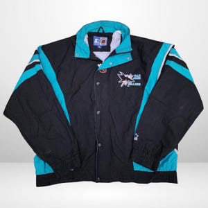 May include: A black and teal San Jose Sharks Starter jacket featuring a high collar, a front button closure, and the team logo embroidered on the chest. The jacket includes long sleeves with elastic cuffs and a white interior lining.