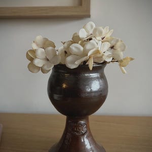 May include: A dark brown ceramic goblet-shaped vase with a rounded bowl and a flared base. The vase features a circular embossed stamp on the stem and is filled with a cluster of off-white, delicate artificial flowers with soft, rounded petals.