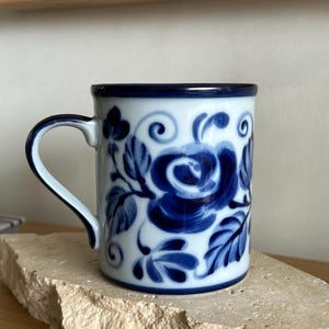 May include: A white ceramic mug featuring a hand-painted dark blue floral pattern with swirling leaves and rose-like blossoms. The mug has a cylindrical body, a curved handle, and a solid dark blue rim along the top edge of the vessel.