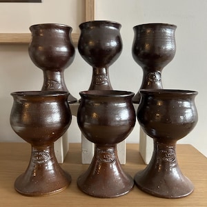 May include: A set of six dark brown glazed ceramic goblets with a rounded bowl and a flared base. Each goblet features a circular stamp on the stem that reads "Studio Pottery W.A. Australia" in raised lettering, indicating their origin and craft.