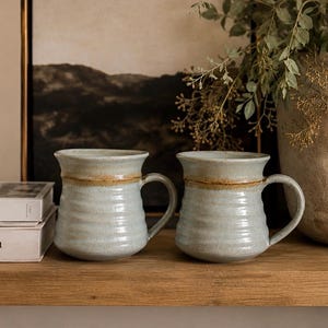 May include: Two ceramic mugs with a light blue, rippled texture and a smooth, tan-coloured band around the upper rim. Each mug features a curved handle on the side and a wide, rounded base, showcasing a handcrafted appearance with subtle colour variations.