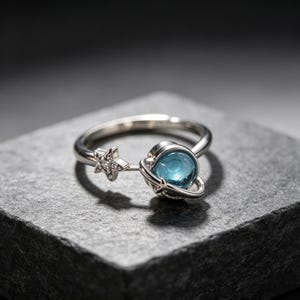 May include: A silver-toned ring featuring a celestial design with a small star encrusted with tiny clear stones and a central blue spherical element surrounded by a thin metal ring, resembling a planet with a ring system in orbit.