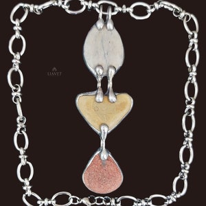 May include: Silver-toned necklace with a pendant featuring three textured stone shapes: a light gray oval, a yellow-beige heart, and a reddish-brown teardrop. The pendant is suspended from a chain-link necklace.