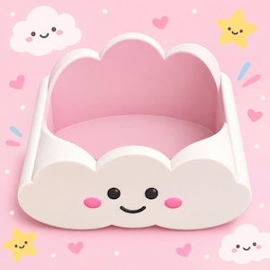 Puede incluir: Un recipiente de almacenamiento con forma de nube blanca, acabado liso e interior rosa. La parte frontal presenta una cara impresa con dos ojos negros circulares, una boca negra curvada y dos pequeñas mejillas ovaladas de color rosa.