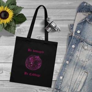 May include: A black tote bag featuring a graphic of a purple cabbage in the center. Above the cabbage, the text reads Be damaged, and below the cabbage, the text reads Be Cabbage. The text and the cabbage graphic are both in a dark pink color.
