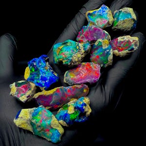 May include: A collection of raw, unpolished opal stones featuring vibrant flashes of iridescent blue, green, and red colors. Each stone displays a unique, jagged texture with natural earthy inclusions, showcasing a high degree of color play.