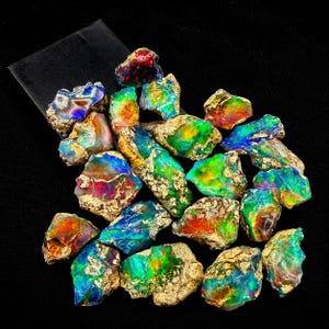 May include: A collection of raw, unpolished opal stones featuring vibrant flashes of iridescent green, blue, red, and yellow colors. Each stone displays a rough, textured matrix surface with natural mineral deposits and irregular, jagged edges.