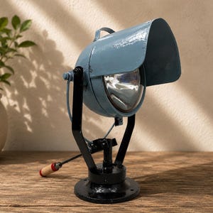 May include: A blue metal spotlight with a large hood and a clear glass lens. The light is mounted on a black metal swivel base with a black support frame. A small handle with a wooden grip extends from the side for adjusting the angle of the light.