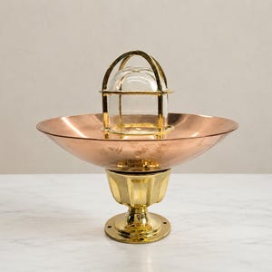 May include: A nautical-style light fixture featuring a polished brass base and a wide, shallow copper bowl. A clear glass dome is protected by a brass cage structure centered within the bowl, creating a metallic, multi-toned lighting piece.