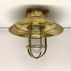 May include: A nautical-style ceiling light fixture crafted from polished brass, featuring a wide circular reflector shade and a protective brass cage surrounding a clear glass cylindrical bulb housing, mounted on a square brass base plate.