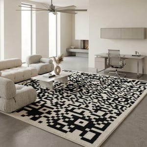 Può includere: Un grande tappeto rettangolare caratterizzato da un motivo geometrico bianco e nero ad alto contrasto che ricorda un codice QR. Il tappeto è posizionato su un pavimento grigio liscio, estendendosi sotto un divano modulare chiaro e un tavolino.