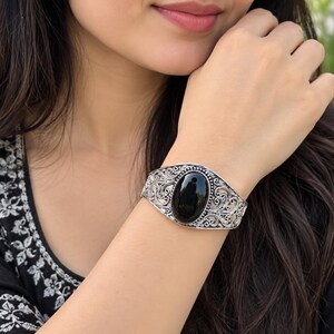 Peut inclure: Un bracelet manchette en argent orné d'une pierre noire de forme ovale en son centre. La bande métallique est décorée de motifs en filigrane complexes et tourbillonnants qui s'étendent sur toute la surface, entourant la pierre polie.