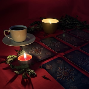 Puede incluir: Una taza y plato de cer&aacute;mica blanca con un l&iacute;quido oscuro, junto a una vela de t&eacute; encendida, una vela grande y varias cartas rectangulares con un dise&ntilde;o de sol dorado sobre fondo azul oscuro, todo sobre una superficie de color rojo.