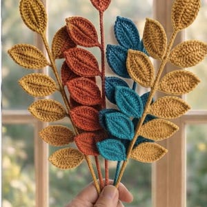 May include: A collection of three crocheted leaf stems featuring textured, pointed leaves in shades of mustard yellow, burnt orange, and teal blue. Each stem is crafted with individual yarn-stitched leaves attached to a central vertical stalk.