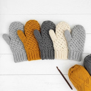May include: Five crocheted mittens in light gray, mustard yellow, dark charcoal gray, cream, and medium gray, each featuring a textured stitch pattern and a ribbed cuff, accompanied by a brown wooden crochet hook and a ball of mustard yellow yarn.