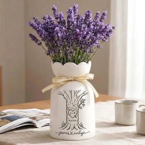 May include: A white ceramic vase with a scalloped rim and a beige ribbon tied around the neck. The vase features a black line drawing of a tree with a heart carved into the trunk containing the text J + E and 05.10.2026, plus the name James Evelyn.