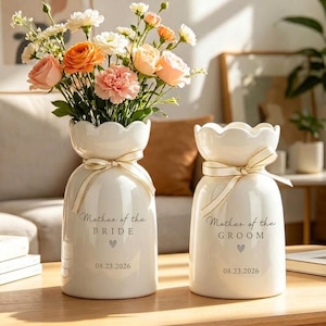 May include: Two white ceramic vases with scalloped rims, each tied with a cream ribbon. One vase is printed with "Mother of the BRIDE" and "08.23.2026", the other with "Mother of the GROOM" and "08.23.2026". Both feature a small grey heart icon.