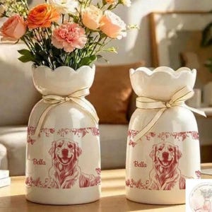 May include: Two cream-colored ceramic vases with scalloped rims and cream ribbons tied around the necks. Each vase features a red line-art illustration of a Golden Retriever dog, the name Bella, and floral accents around the top and bottom.