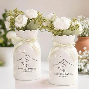 May include: Two white ceramic vases with scalloped rims, each tied with a cream ribbon. The vases feature a black line drawing of a mountain path with two figures and the text "SOPHIA + DANIEL 10.20.2025". Each vase holds a bouquet of white flowers.